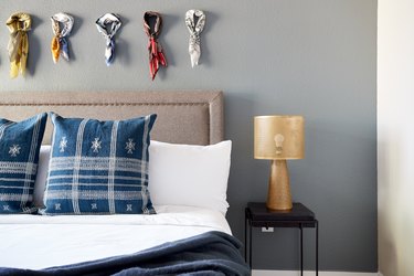 Eclectic bedroom with gray upholstered headboard, blue pillows, gray wall, brass lamp, nightstand.